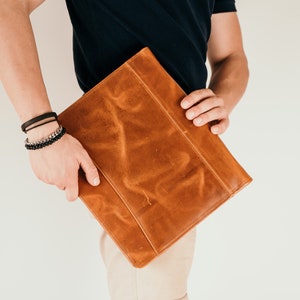 May include: A brown leather portfolio case with a flap closure. The case is held by a person wearing a black shirt and beige pants.