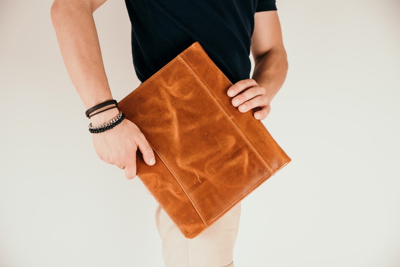 May include: A brown leather portfolio case with a flap closure. The case is held by a person wearing a black shirt and beige pants.