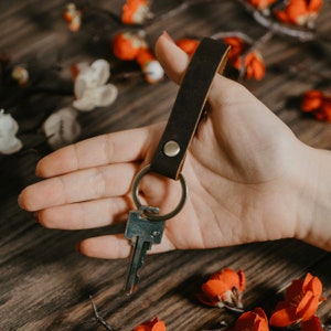 May include: A brown leather keychain with a silver key ring and a single key attached. The keychain is held in a hand against a wooden background.