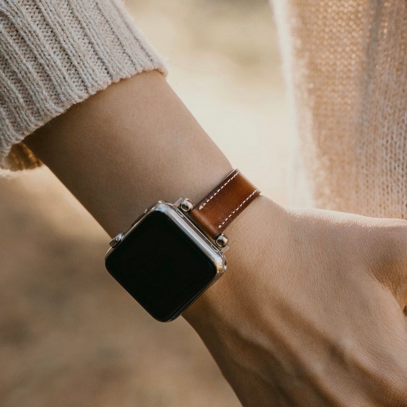 May include: A silver Apple Watch with a brown leather band. The watch face is black and the band has white stitching.