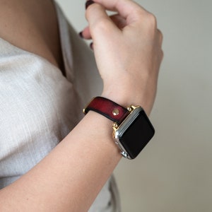Op de afbeelding: Een rood leren horlogebandje met gouden accenten en een zilveren smartwatch met een zwart scherm.