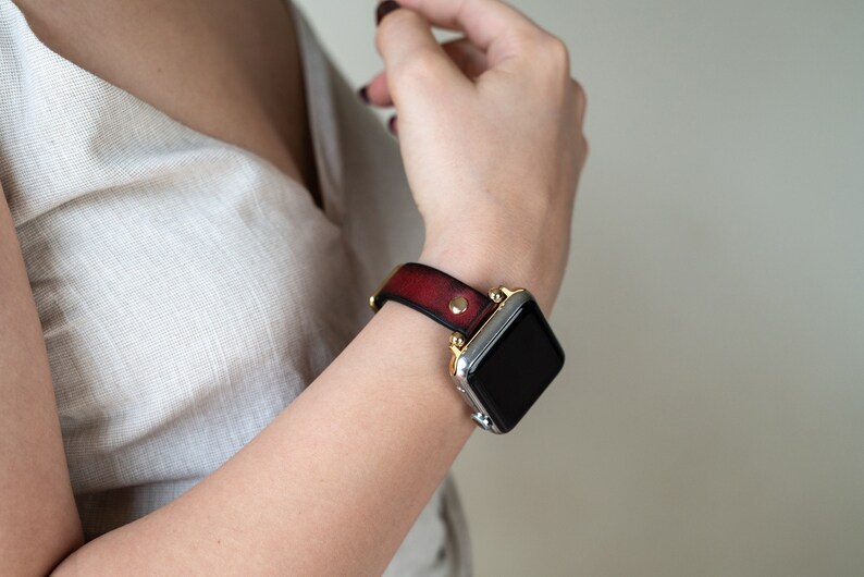 Op de afbeelding: Een rood leren horlogebandje met gouden accenten en een zilveren smartwatch met een zwart scherm.