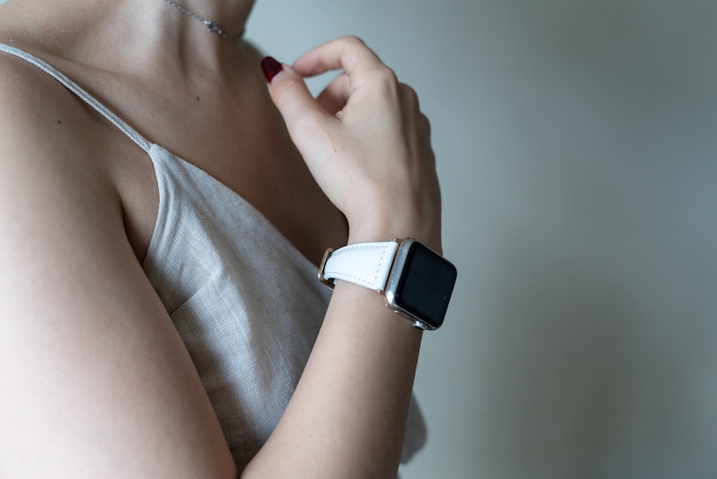 May include: A white leather watch band with a silver watch face. The watch is on a person's wrist.