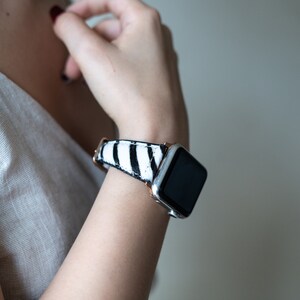 May include: A white and black striped watch band with a rose gold buckle. The watch face is black and the watch case is silver.