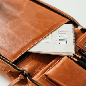 May include: A brown leather portfolio case with a zipper closure. The case is open and contains a notebook, pens, and a card holder. The notebook has the text "lutart" printed on the cover.
