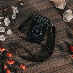 May include: A black and silver medium format camera with a brown leather strap, surrounded by white and orange blossoms on a wooden surface.