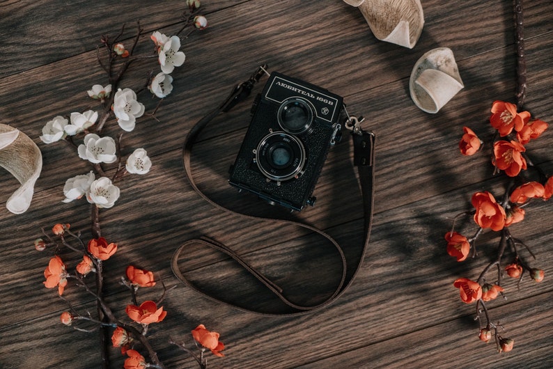 May include: A black and silver medium format camera with a brown leather strap, surrounded by white and orange blossoms on a wooden surface.