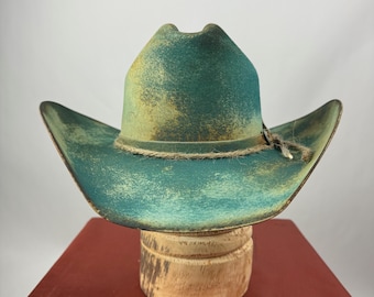 May include: A distressed turquoise cowboy hat with a weathered appearance. The hat features a wide brim and a tall crown, with a rope band around the base. The hat is displayed on a wooden hat stand, set against a red surface.