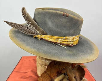 May include: A gray felt cowboy hat with a yellow bandana and brown feathers. The hat is sitting on a wooden stand with dried leaves and grasses.
