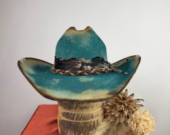 May include: A teal cowboy hat with a distressed finish and dark brown edges. The hat features a decorative band composed of a dark, textured leather-like strip layered with a braided brown cord, creating a rugged and layered appearance.