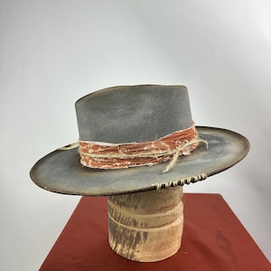 May include: A grey felt cowboy hat with a wide brim and a distressed look. The hat has a brown leather band with a red and white fabric band wrapped around it. The hat is sitting on a wooden hat stand.