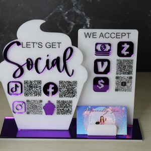 May include: White cupcake-shaped sign with the text "Let's Get Social" and QR codes for social media platforms. A separate white sign reads "We Accept" with purple icons for various payment methods. A business card with a pink background and the text "Personally You" is displayed in a holder.