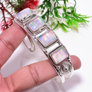 Puede incluir: Una pulsera de plata con cinco piedras de ópalo rectangulares. Los ópalos muestran un juego de colores con tonos iridiscentes de rosa, verde y azul. La pulsera tiene un diseño detallado y texturizado alrededor de las piedras y un adorno en espiral.