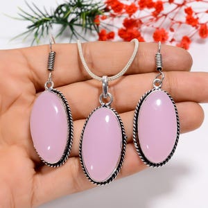 May include: A jewelry set featuring a pendant necklace and matching earrings. The set includes a silver-toned chain and earring hooks. Each piece showcases a large, oval, light pink stone with a decorative, dark silver rope-like border. The jewelry is displayed on a hand.