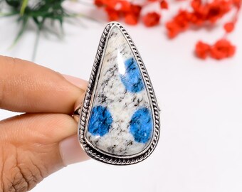 May include: A teardrop-shaped ring featuring a large, polished k2 stone with a white base and blue spots. The stone is set in a silver-colored metal with a textured border. The ring is held by a hand, showcasing the jewelry's design and color.