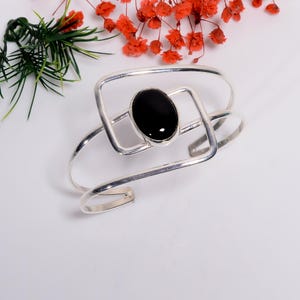 May include: A silver-toned cuff bracelet with a large, oval black stone at the center. The bracelet has a modern, open design with geometric shapes. The background includes sprigs of green and red flowers.