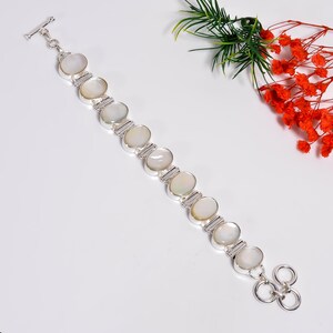 May include: A silver bracelet featuring oval, iridescent gemstones. The bracelet has a toggle clasp and is accented with rectangular silver links. The gemstones are set in a silver bezel setting. The bracelet is displayed on a white surface with greenery and red flowers.
