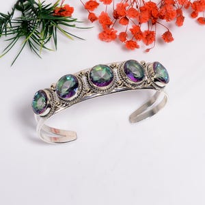 Puede incluir: Una pulsera de plata con cinco piedras preciosas ovaladas e iridiscentes. Las piedras están engastadas en un bisel decorativo en forma de cuerda. La pulsera tiene un diseño abierto, con los extremos ligeramente curvados.