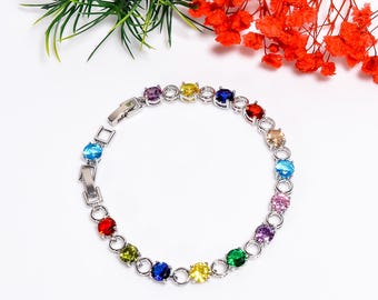 Può includere: Un bracciale in argento con una serie di pietre preziose colorate. Le pietre preziose sono rotonde e incastonate in una catena d'argento. I colori includono rosso, arancione, giallo, verde, blu, viola e rosa. Il bracciale è esposto su una superficie bianca.