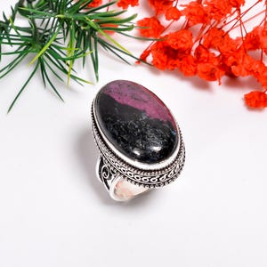 Könnte beinhalten: Silberring mit einem ovalen Edelstein mit einem schwarz-rosa Muster. Der Ring hat eine detaillierte, dekorative Fassung. Im Hintergrund sind grüne und rote florale Elemente zu sehen.