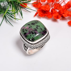 May include: A silver ring with a large, square-cut gemstone. The gemstone is a mix of green, black, and purple. The ring has a detailed, patterned band. The background includes green leaves and red flowers.