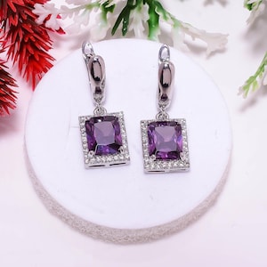 May include: A pair of silver-toned drop earrings featuring a rectangular, deep purple faceted gemstone at the center. Each stone is surrounded by a border of small, clear white stones set in a metallic frame with a secure hinged lever-back closure.