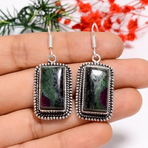 May include: Silver earrings featuring rectangular pendants with green and red gemstones. The pendants are set within a beaded frame and hang from coiled ear wires. The earrings are shown against a neutral backdrop.