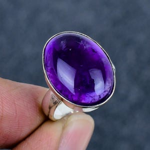 May include: A silver ring with a large, oval amethyst gemstone. The deep purple stone is set in a polished silver band. The ring's design is simple, highlighting the gemstone's beauty. The ring is a statement piece, suitable for everyday wear or special occasions.
