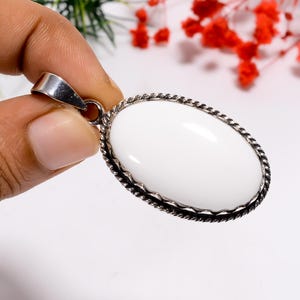 Puede incluir: Colgante ovalado blanco con un borde de cuerda retorcida de color plateado. El colgante est&aacute; sujeto por una fianza de color plateado. El fondo es blanco con algunas flores rojas.