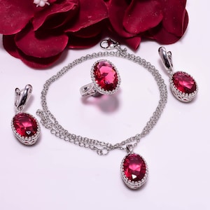 Op de afbeelding: Een sieradenset met een ring, oorbellen, ketting en armband. Elk stuk heeft een zilverkleurige metalen zetting en een grote, ovale, rode edelsteen. De sieraden worden gepresenteerd op een wit oppervlak met rode bloemblaadjes op de achtergrond.