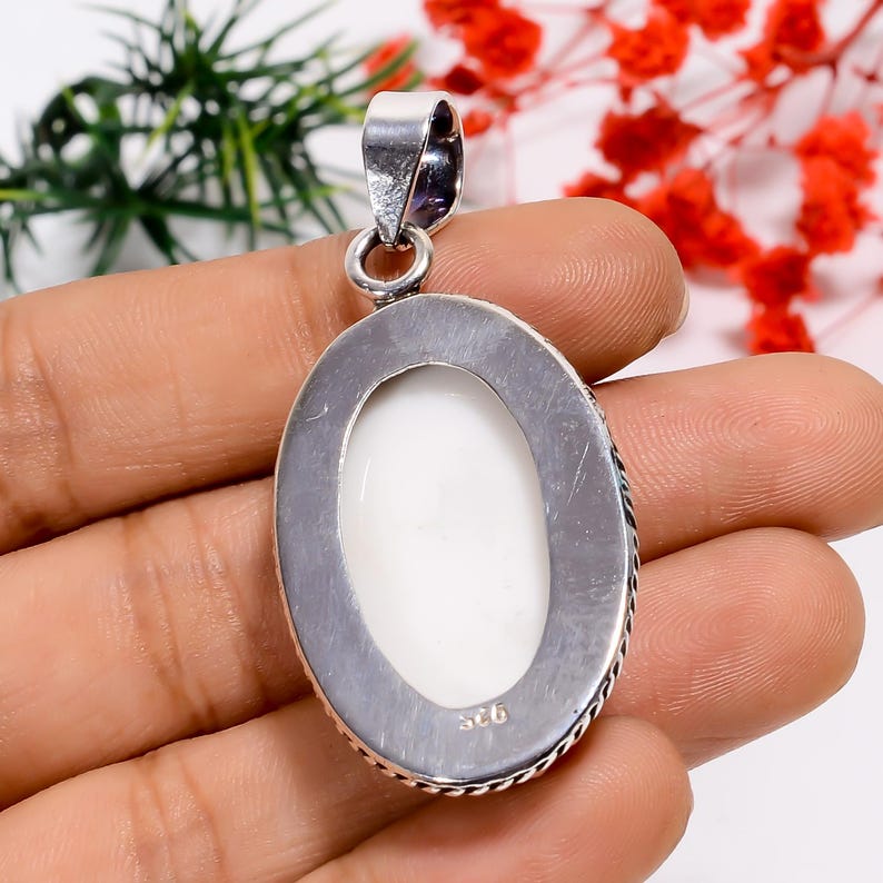 Puede incluir: Colgante ovalado con un centro de piedra blanca, engastado en un marco de metal plateado. El marco tiene un borde en forma de cuerda y un lazo para una cadena. La parte posterior del colgante es visible, con marcas en el metal.