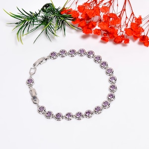Op de afbeelding: Een zilveren armband met een reeks ronde, bezel-set, paarse edelstenen. De armband heeft een eenvoudige sluiting en wordt gepresenteerd op een wit oppervlak, met groene en oranje bloemaccenten op de achtergrond.