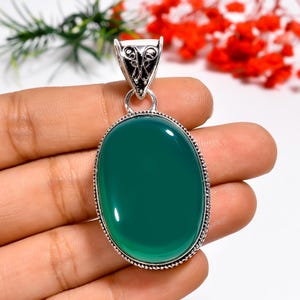 Puede incluir: Colgante ovalado de piedra preciosa verde con una fianza decorativa plateada. El colgante tiene una superficie pulida y brillante y un borde plateado detallado. La piedra preciosa es de un tono verde intenso y rico. El colgante se sostiene en una mano.