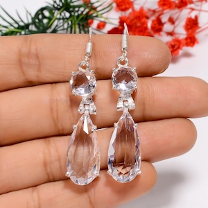 Op de afbeelding: Een paar zilveren oorbellen met heldere, gefacetteerde edelstenen. Elke oorbel heeft een ronde steen aan de bovenkant, verbonden met een druppelvormige steen eronder. De oorbellen worden omhoog gehouden door een zilveren haak.