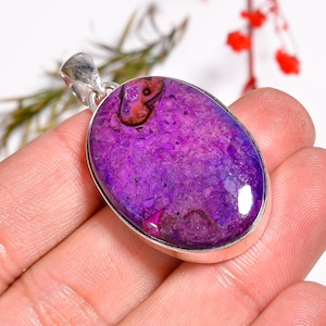 May include: A purple gemstone pendant with a silver bail. The oval-shaped stone has a mottled texture with shades of purple and brown. The pendant is held in a hand against a white background.