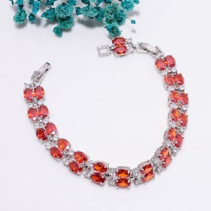 Op de afbeelding: Een zilveren armband met rijen ovale en vierkante oranje edelstenen, afgewisseld met kleinere heldere stenen. De armband heeft een veilige sluiting en wordt gepresenteerd op een witte ondergrond.