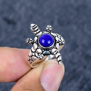 Può includere: Anello color argento a forma di tartaruga, con una grande pietra rotonda blu scuro al centro. Il guscio e gli arti della tartaruga presentano motivi strutturati. L'anello è tenuto da una persona.