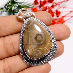 May include: A teardrop-shaped pendant with a silver-colored bail. The pendant features a polished, light brown stone with a unique, patterned surface. The stone is set within a textured, silver-colored frame, creating a striking contrast.