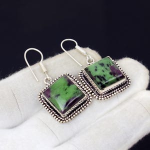 May include: A pair of silver-toned earrings with square, green and purple gemstones. The gemstones are set in a decorative, beaded frame. The earrings have a simple hook design.