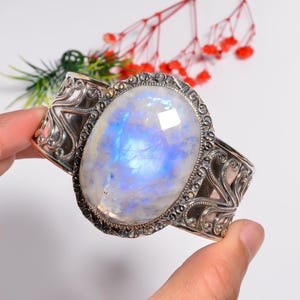 May include: A silver cuff bracelet with an oval moonstone. The stone has a blue and white glow. The bracelet has an ornate, engraved design on the band and around the stone setting. The bracelet is held by a person.