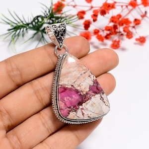 Puede incluir: Un colgante con una piedra triangular grande con una mezcla de colores rosa y crema. La piedra está engastada en un marco de metal plateado con detalles intrincados en la parte superior. El colgante se sujeta con una argolla plateada.