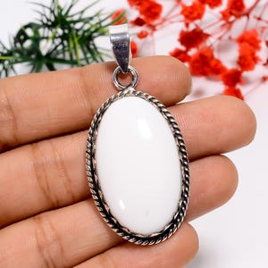 Puede incluir: Colgante ovalado blanco con una fianza plateada y un borde retorcido en forma de cuerda. El colgante se sostiene en una mano sobre un fondo de follaje verde y rojo.