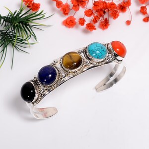 Può includere: Bracciale a polsino in argento con cinque pietre preziose colorate: nero, blu, marrone, turchese e rosso. Le pietre sono incastonate in montature d'argento ornate. Il bracciale aperto è appoggiato su una superficie bianca.