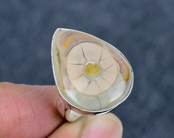 Könnte beinhalten: Silberring mit einem tropfenförmigen Stein. Der Stein hat ein cremefarbenes, gelbes und beiges Muster mit einem zentralen Design, das einer Blume oder einem Auge ähnelt. Das Design hat strahlenförmige Linien.