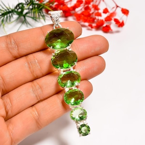 May include: A silver pendant featuring a series of oval-cut, vibrant green gemstones. The gemstones are arranged in a vertical line, creating a striking and elegant design. The pendant is held in a hand, showcasing its size and detail.