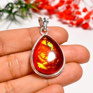 May include: A teardrop-shaped pendant with a vibrant, iridescent gemstone. The gem displays fiery red, orange, and yellow hues, set in a silver-colored metal frame. The pendant is suspended from a matching bail, showcasing a unique and eye-catching design.