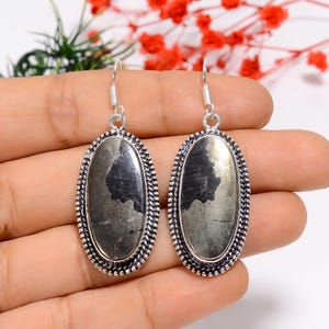 Può includere: Un paio di orecchini pendenti color argento. Ogni orecchino presenta una pietra ovale grigio scuro con una superficie strutturata, incorniciata da un bordo decorativo argentato. Gli orecchini sono tenuti in una mano, con uno sfondo sfocato di fiori rossi.