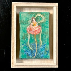 May include: A framed artwork featuring a woman in traditional attire seated on a large pink flower. The background is a textured teal with blue floral patterns. The artwork is encased in a light-colored wooden frame.