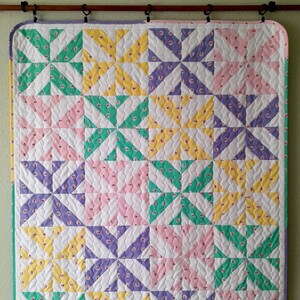 May include: A quilted baby blanket with a patchwork design featuring a repeating pattern of green, yellow, pink, and purple pinwheels. The blanket is made of cotton fabric and has a white background.