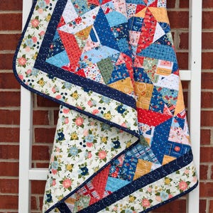 Op de afbeelding: Een patchwork quilt met een verscheidenheid aan bloemrijke en geometrische stoffen patronen in tinten rood, blauw, geel en wit. De quilt heeft een donkerblauwe rand met een golvend stiksel en een witte bloemrijke stoffen rand aan de buitenkant.